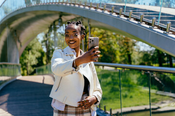 Obraz na płótnie Canvas African woman making selfie photo while walking and admiring of a modern metal pedestrian bridge and looking at city park. A black woman stands at the bridge and takes a selfie on a smartphone.