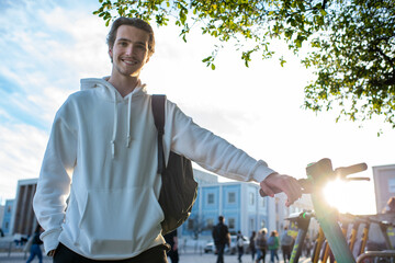 Student smiling while holding onto an electric scooter. Happy man standing close to an environmentally friendly vehicle. A cheerful young man with a backpack outdoors about to take a ride on a scooter