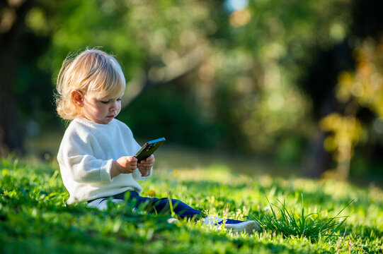 Kid Holding A Phone In His Hands While Outdoors. A Caucasian Child Staring Into A Smartphone With Interest, Sitting In A Park Alone On A Sunny Day. Cute Child Sitting In A Park On A Nice Sunny Day.