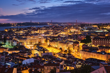 Fototapeta premium Night view of Lisbon famous view from Miradouro da Senhora do Monte tourist viewpoint of Alfama and Mauraria old city districts, 25th of April Bridge in the evening twilight. Lisbon, Portugal
