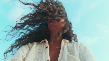 Young cute positive Indian woman with smile holding camera and making funny grimace blowing hair off face and walking along sandy shore dressed in casual summer shirt located on beach of sea island.