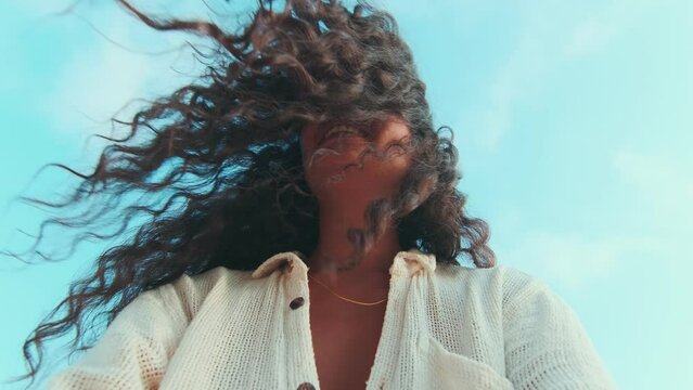 Young cute positive Indian woman with smile holding camera and making funny grimace blowing hair off face and walking along sandy shore dressed in casual summer shirt located on beach of sea island.