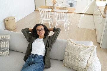 Relaxed girl in glasses dreams on cozy couch at home. Calm woman enjoys lazy break, resting with hands behind head.