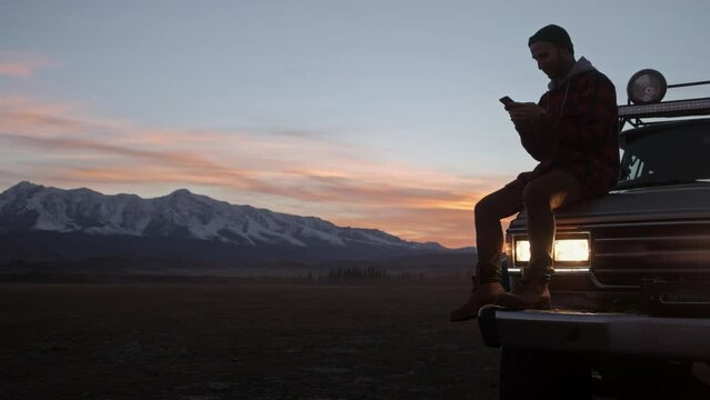 Male traveler is making route, while is sitting on a car hood against mountains view. Caucasian man, traveler and adventurer with a beard consulting his mobile phone leaning on his 4x4 car