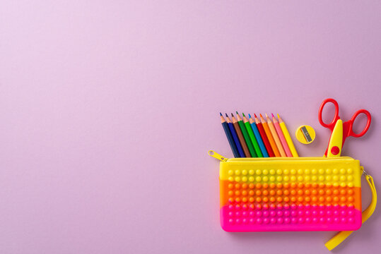 Artistic classroom setup. Top view of range of colorful items: pop-it pencil case with vibrant pencils, cutting scissors, sharpener on lilac backdrop, with empty space for text or promotional content