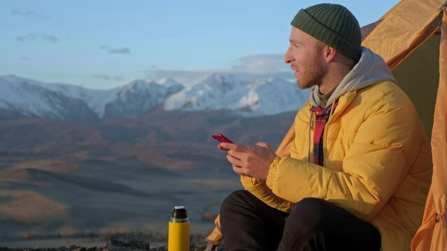 Traveler male uses mobile phone sitting in tent in wild nature. Close up image man hands with mobile phone. Bearded Man portrait using his mobile phone and sitting in camping tent in wild nature