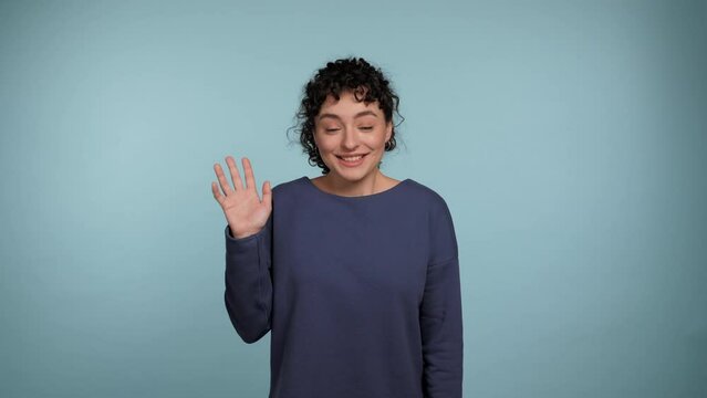Smiling young curly woman greets by waving her hand, showing hello gesture looks into camera. Portrait positive female standing on isolated light blue background. People emotions lifestyle concept