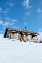 Man walk up steep ski slope to mountain cabin with help of ski poles, carry backpack with snowboard attached. Get ready for downhill on fresh powder snow. Ski or snowboard randoneering