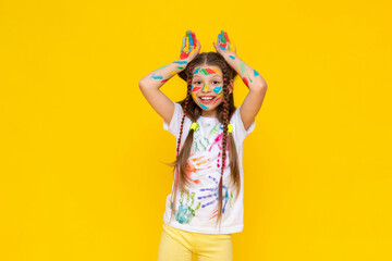 Portrait of a girl with a face painted with multicolored paints. Children's creativity. The child shows his ears above his head. Yellow isolated background.