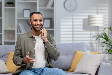 Portrait of a young Hispanic man sitting on the couch at home and talking on the phone with a smile