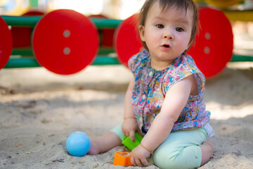 Fototapeta na wymiar Adorable 9 months old baby playing outdoors - lifestyle portrait of mixed ethnicity Asian Caucasian baby girl playing with block toys happy and carefree at playground sitting on sand