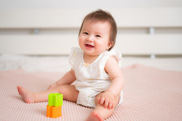 lifestyle home portrait of 9 months old mixed ethnicity Asian Caucasian baby girl playing happy and carefree on bed crawling and handling color blocks in childhood and nursery concept