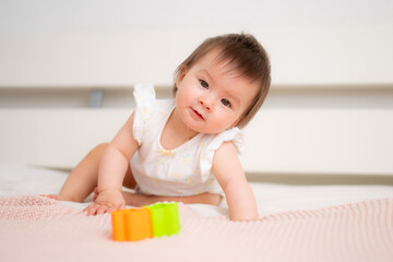 lifestyle home portrait of 9 months old mixed ethnicity Asian Caucasian baby girl playing happy and carefree on bed crawling and handling color blocks in childhood and nursery concept