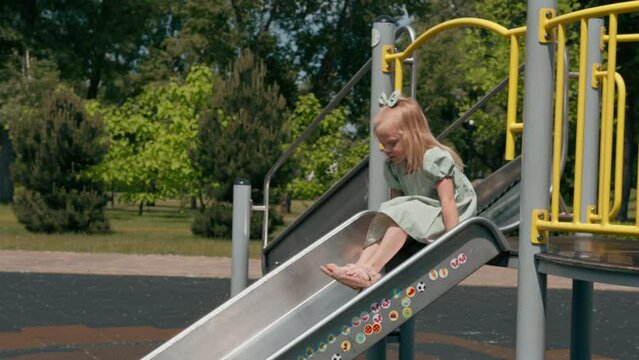 A cute little girl in a green summer dress with a bow on the playground in the kindergarten is having fun climbing down the slide childhood