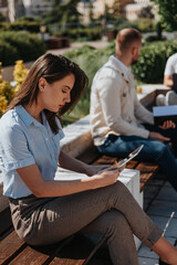 Brunette girl working remotely on tablet while sitting on a bench in the city