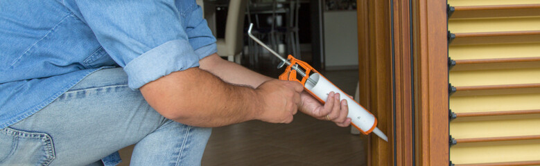Image of the hands of a handyman who with a silicone gun installs and seals doors and windows against drafts. Home thermal insulation improvements. Horizontal banner 
