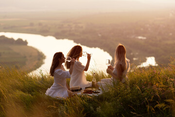 Friends having picnic on the hill at sunset. Three girlfriends eating and drinking wine on outdoor...