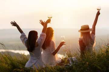 Back view of women enjoying hills view and drinking wine. Happy people spending time picnic leisure together on the sunset.