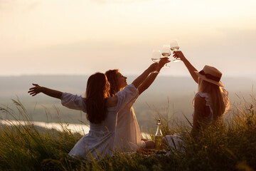 Three young beautiful happy woman is resting at picnic at sunset. Girlfriends enjoy sun day evening on beautiful green meadow. Vacation, picnic, friendship or holiday concept.