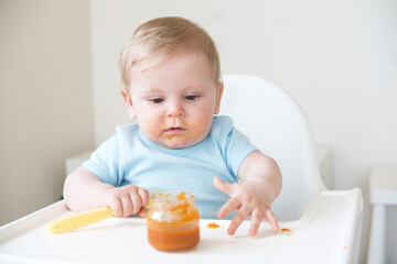 dirty baby boy holding spoon eating bland mashed food sitting, on high chair. first baby food
