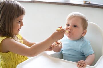 baby boy eating bland mashed food sitting on high chair, older sister feeding child 