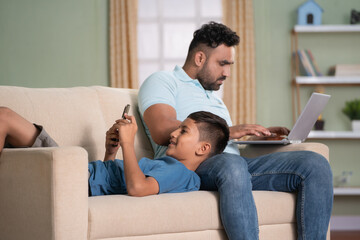 Fototapeta premium Indian father working on laptop while son using mobile phone by sleeping on lap on sofa at home - concept of Digital connection, technology addiction and cyberspace.