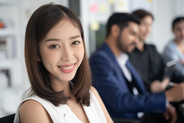 Attractive asian young confident businesswoman sitting at the office table with group of colleagues in the background. Business woman with coworkers in meeting room.