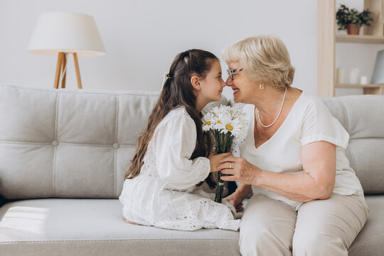 I Love You, Granny. Portrait Of Cute Little Girl Embracing And Kissing Her Grandmother In Cheek Giving Bunch Of Flowers And Postcard, Greeting Woman With Holiday, Women's Or Mother's Day Or Birthday.