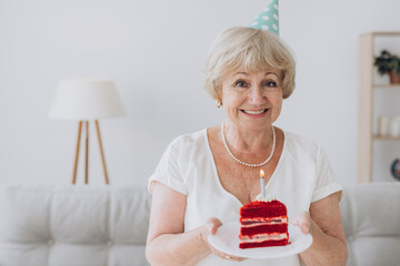 Happy senior woman celebrating her birthday wearing a party hat, holding a cake with a candle - Powered by Adobe