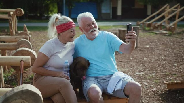 Retired smiling couple and their pet dog take selfies photos with phone after training in an eco-gym in nature