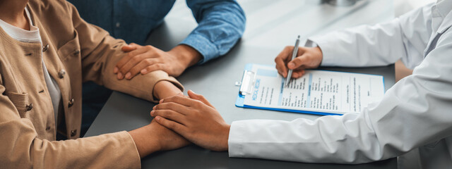 Doctor providing compassionate healthcare consultation while young couple patient holding hand, comfort each other after infertile report. Reproductive and medical fertility consulting. Neoteric