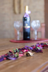 path of colored petals in focus, in the background a bottle of wine with two glasses, on a wooden table