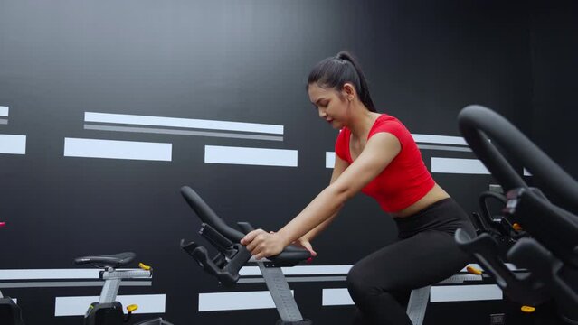 Young asian woman riding bicycle machine for workout training in fitness gym club, female cycling for exercise with motivation and concentration for wellbeing and weightloss, sport and healthcare.