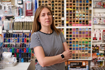 Confident young woman, sales manager, seller looking at camera, standing with her arms folded in school stationery shop