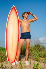 Male teenage surfer standing with his surfboard while looking out at the ocean for waves 