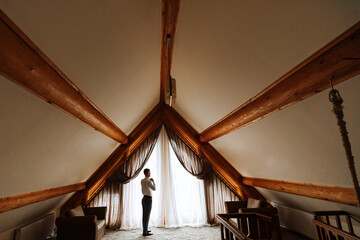 A man is standing by the window in a hotel room. Wide Plan photo. The groom dresses and prepares for the wedding. A wonderful interior for relaxation