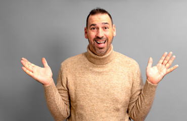 Bearded Hispanic man in his 40s wearing a turtleneck sweater with arms outstretched in an expression of surprise, isolated on gray studio background.