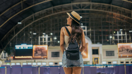 Rear view of young female traveller standing with backpack and hat at train station looking around...