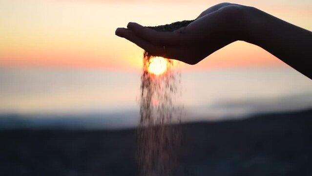 Close up of woman pouring sand running through fingers slow motion at the beach with sun flare and blue sky. Beautiful scene of young lady playing with sand on sunrise or sunset.
