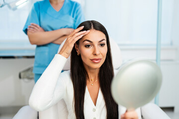 Beautiful and happy young woman sitting in medical chair and looking in the mirror. She is...