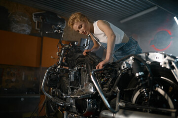 Naklejka na ściany i meble Beautiful woman mechanic posing for camera nearby motorcycle at workshop