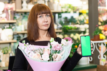 Floristry. Flowers. A florist girl with a bouquet of flowers and a phone with a green screen, a chromakey. Shop on the background of a showcase with fresh flowers and garden decor.