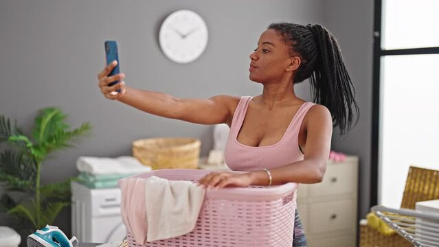 African american woman making selfie by smartphone leaning on basket with clothes at laundry room