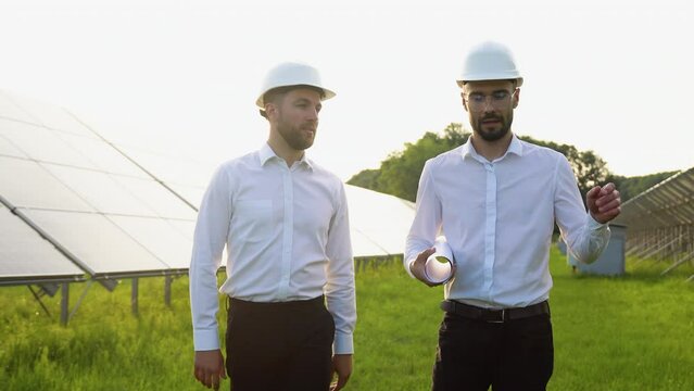 Two investors businessmen walking at solar farm and discussing efficient plan of construction. Two men in protective helmet and uniform talking. Concept of alternative energy