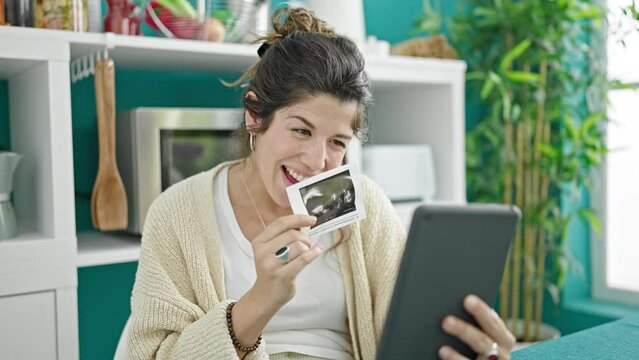 Young pregnant woman having video call showing baby ultrasound at dinning room