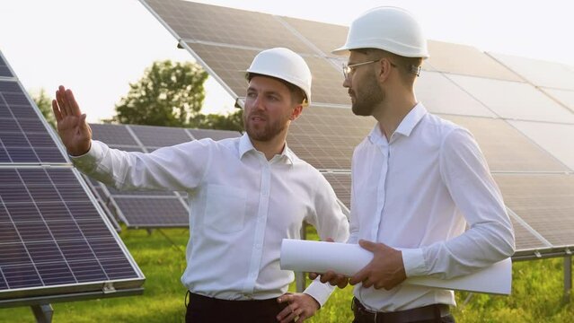Two men in hard helmets discussing project at solar farm. Male engineers with scrolls of engineering drawing communicating. Concept of green energy