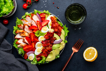 American Cobb salad with grilled chicken, cherry tomatoes, boiled eggs, bacon, avocado and lettuce, black stone table background, top view