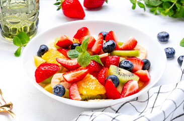 Fototapeta na wymiar Fruit and berry salad with fresh strawberries, blueberries, banana, kiwi, orange and mint leaves, white table background, top view