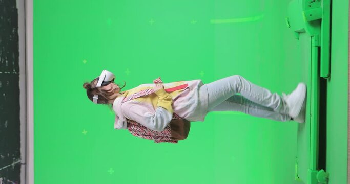 Vertical shot on green screen background. VR or AR online walk. Girl with a backpack in augmented reality headset walks through the virtual world and looks around. Walking on a treadmill side view.