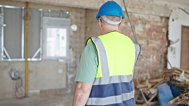 Middle age grey-haired man builder wearing hardhat standing backwards at construction site